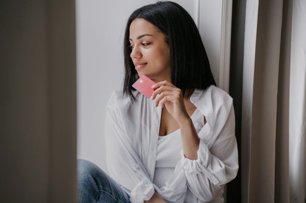 Dreamy African American woman sitting on windowsill looking outside holding credit card, wearing